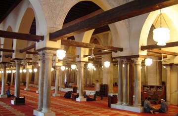 A view from inside of a prayer space with orange carpets spread through out the hall with some people sitting in different corners and praying.